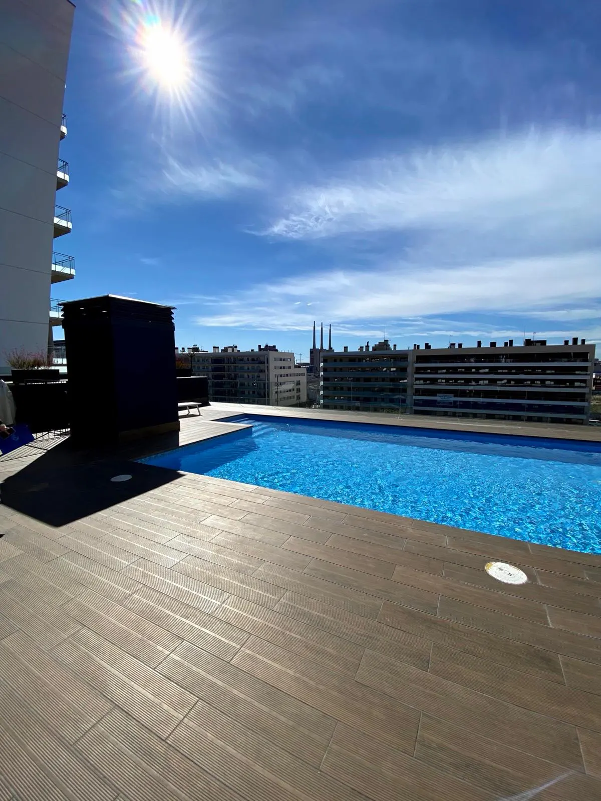 Rooftop infinity pool with blue water and wood-look tile deck under a bright sunny sky with city buildings in the distance.
