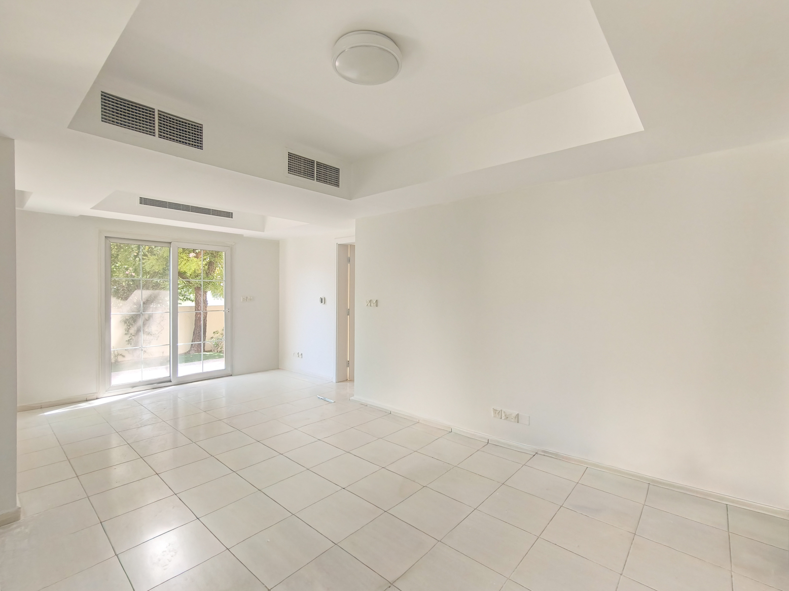 Empty, bright white room with tiled floors, a recessed ceiling with a light fixture, and sliding glass doors to a garden. [AI-generated text]