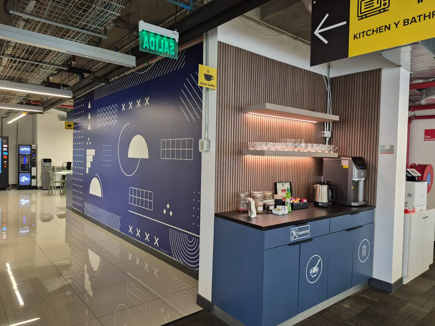 Office break area with blue cabinets, coffee maker, and snacks. A blue wall with white geometric shapes is on the left.