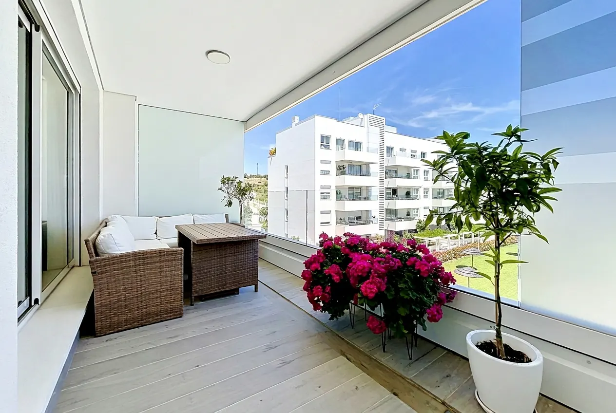 A bright balcony with wicker furniture, pink flowers, and a potted tree overlooks a white building under a blue sky.