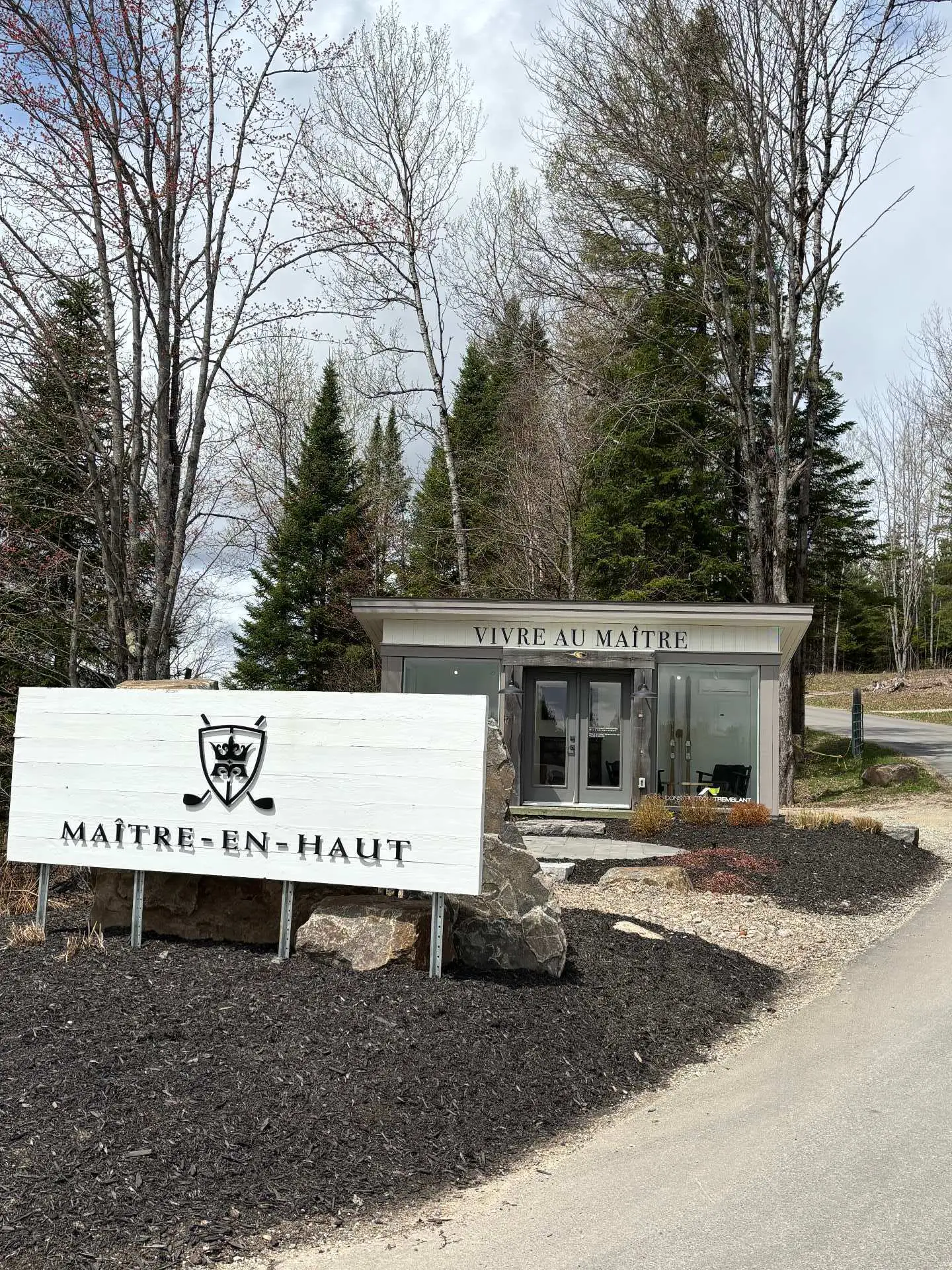 Entrance to Maître-en-Haut with a white sign, logo, and black mulch. A building with "Vivre au Maître" is behind the sign. Trees surround the area.