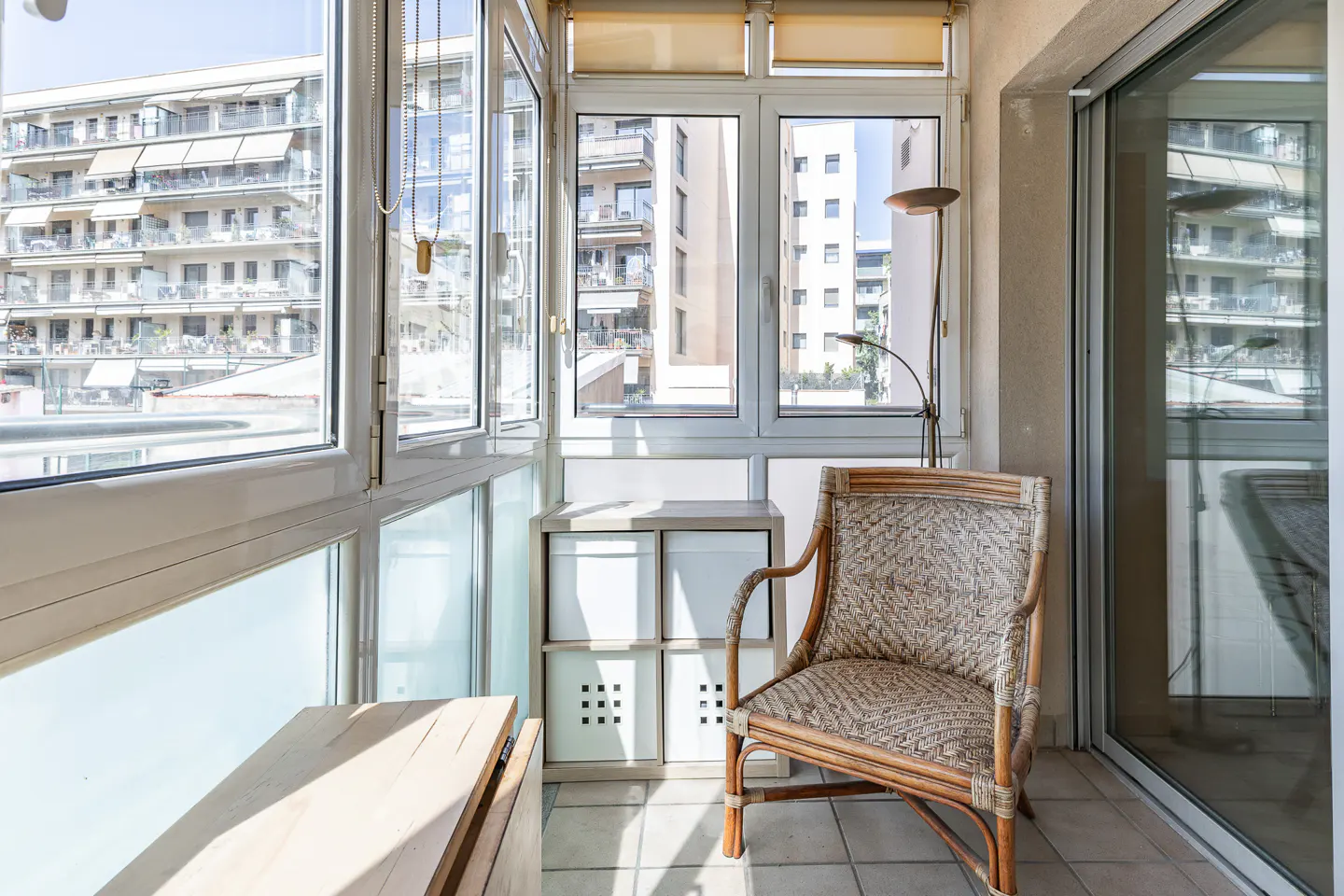 Sunlit balcony with a wicker chair, storage unit, and a view of buildings through white-framed windows.