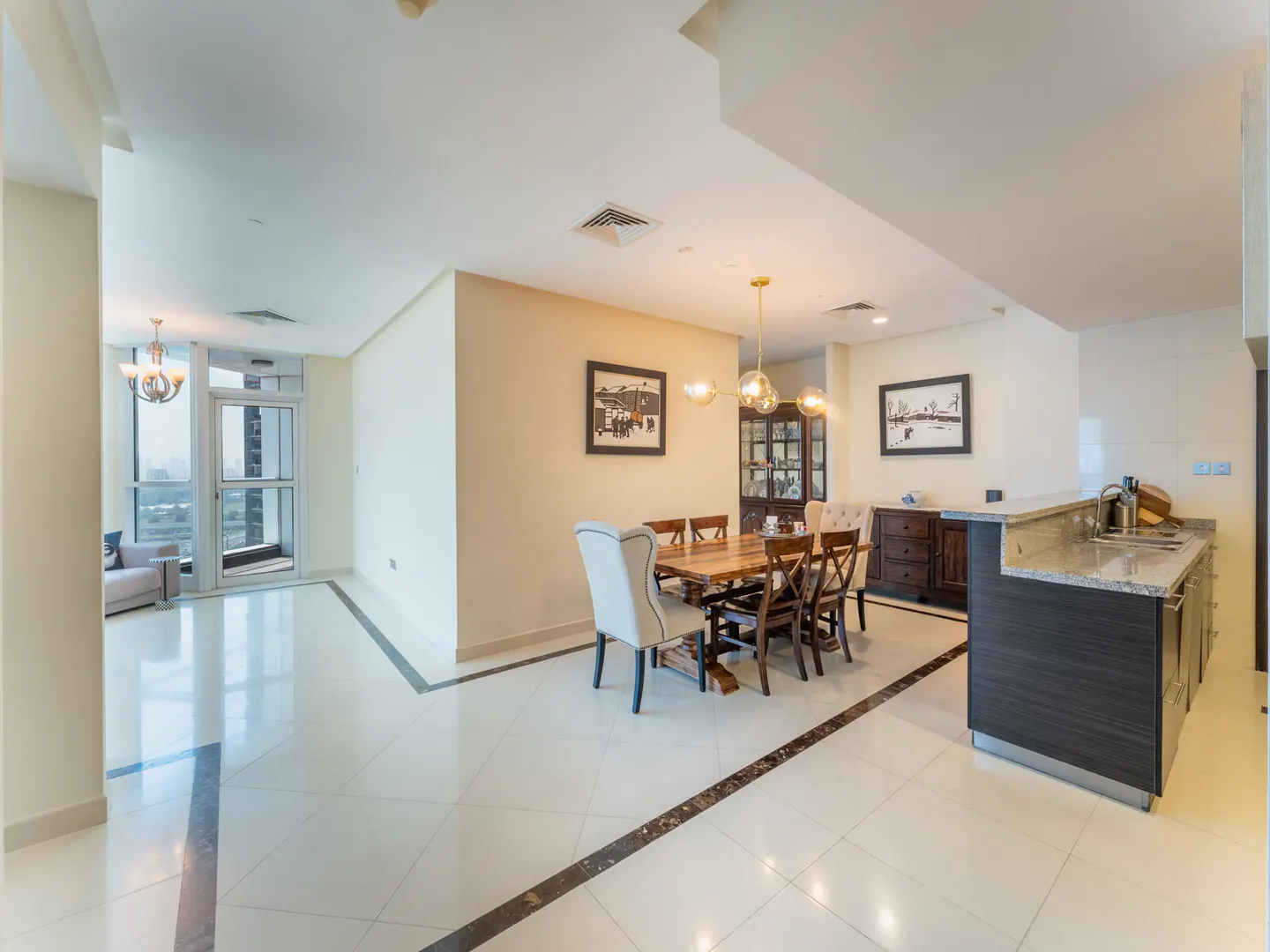 Bright, open-plan apartment with dining table, kitchen counter, and living area with balcony access. Cream walls and white tile floors.