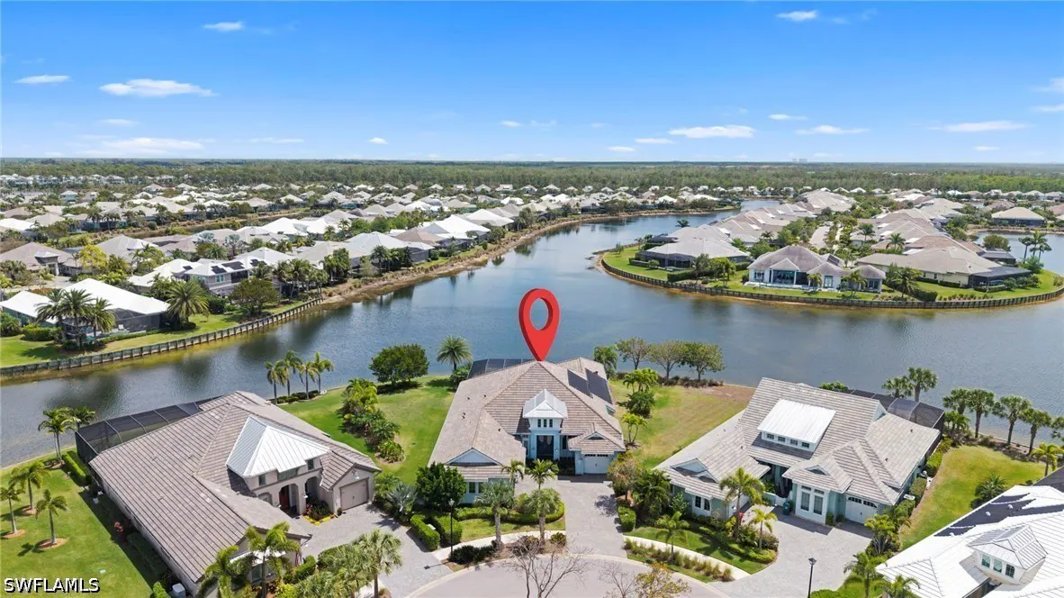 Aerial view of a beige house with a red location pin on the roof, surrounded by water and other houses.