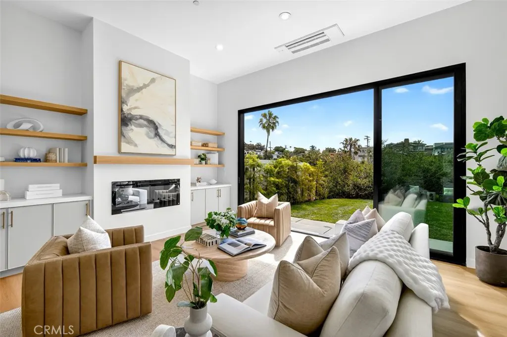 Bright living room with white sofa, tan chairs, and a black-framed window overlooking a green lawn and palm trees. A modern fireplace and shelves add detail.