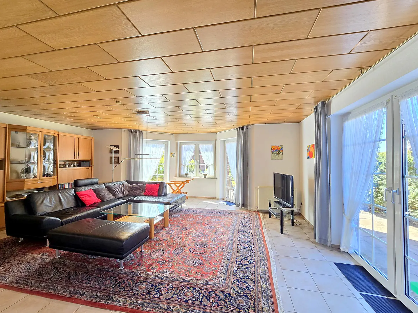 A living room with a black leather sofa, red pillows, a glass table, and a patterned rug. There are windows with white curtains and a wooden ceiling.