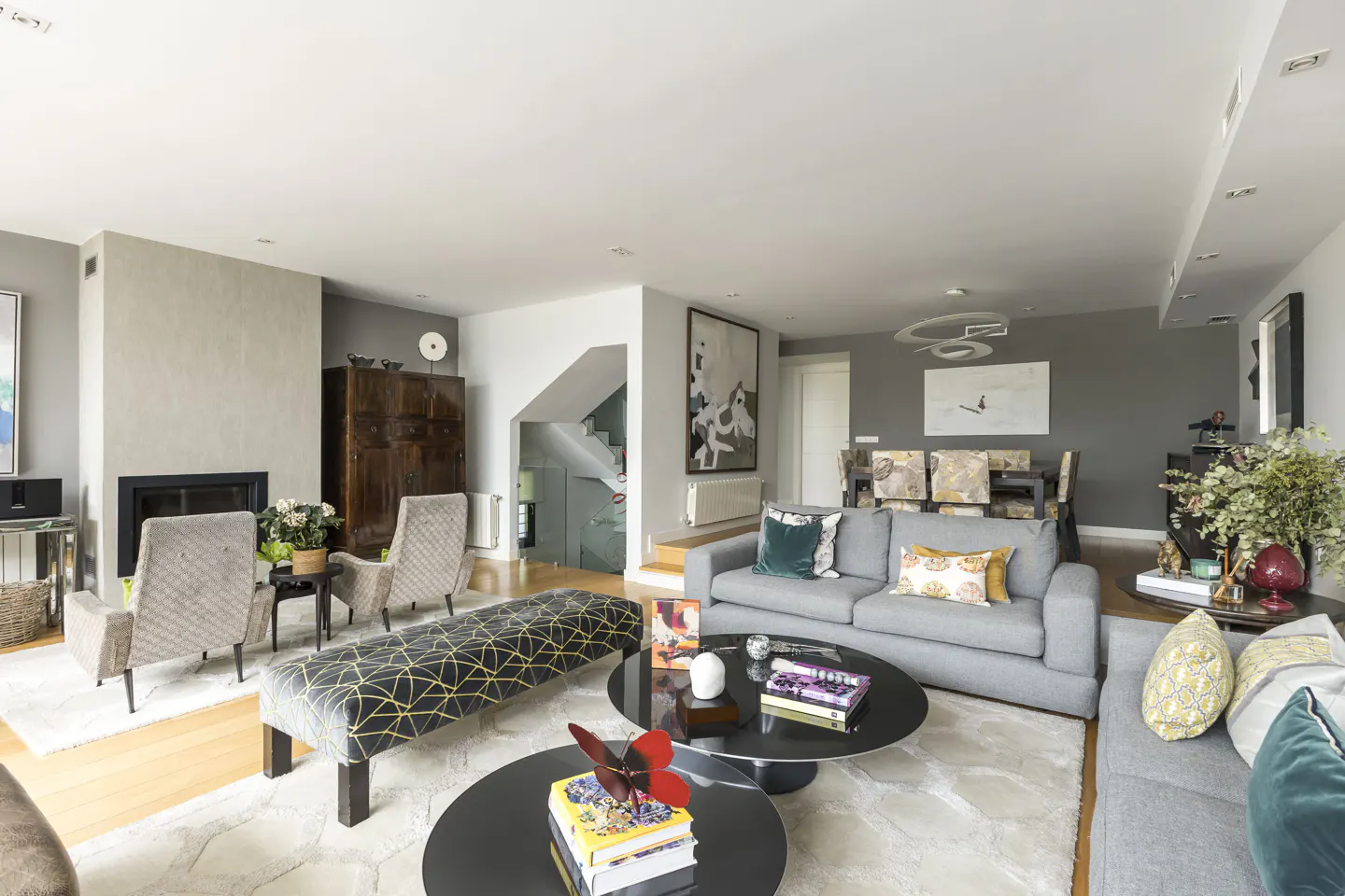 Bright, modern living room with gray sofas, black coffee tables, and a fireplace. Art adorns the walls, and a staircase is visible in the background.