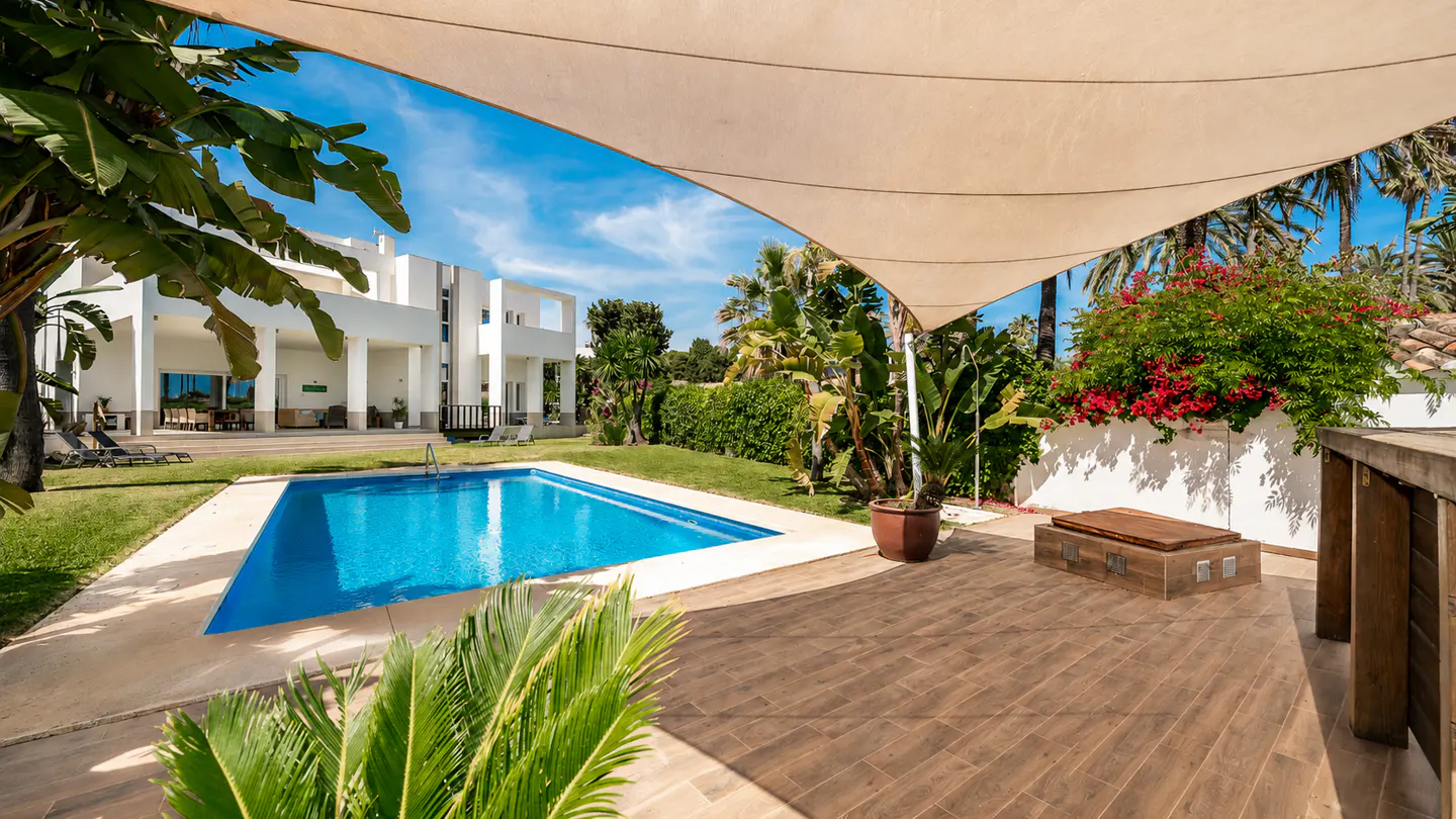 Luxury home exterior with a blue pool, white building, and tropical plants under a beige shade sail.