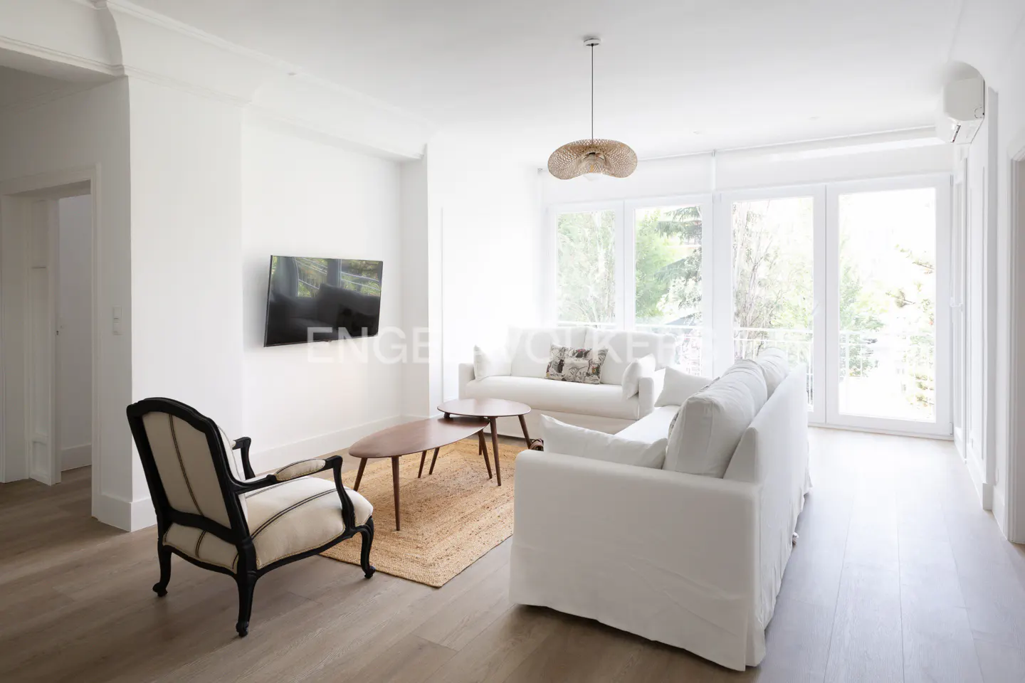 Bright living room with white sofas, wood floors, and a jute rug. A black armchair sits near a TV. Large windows offer a view of trees.