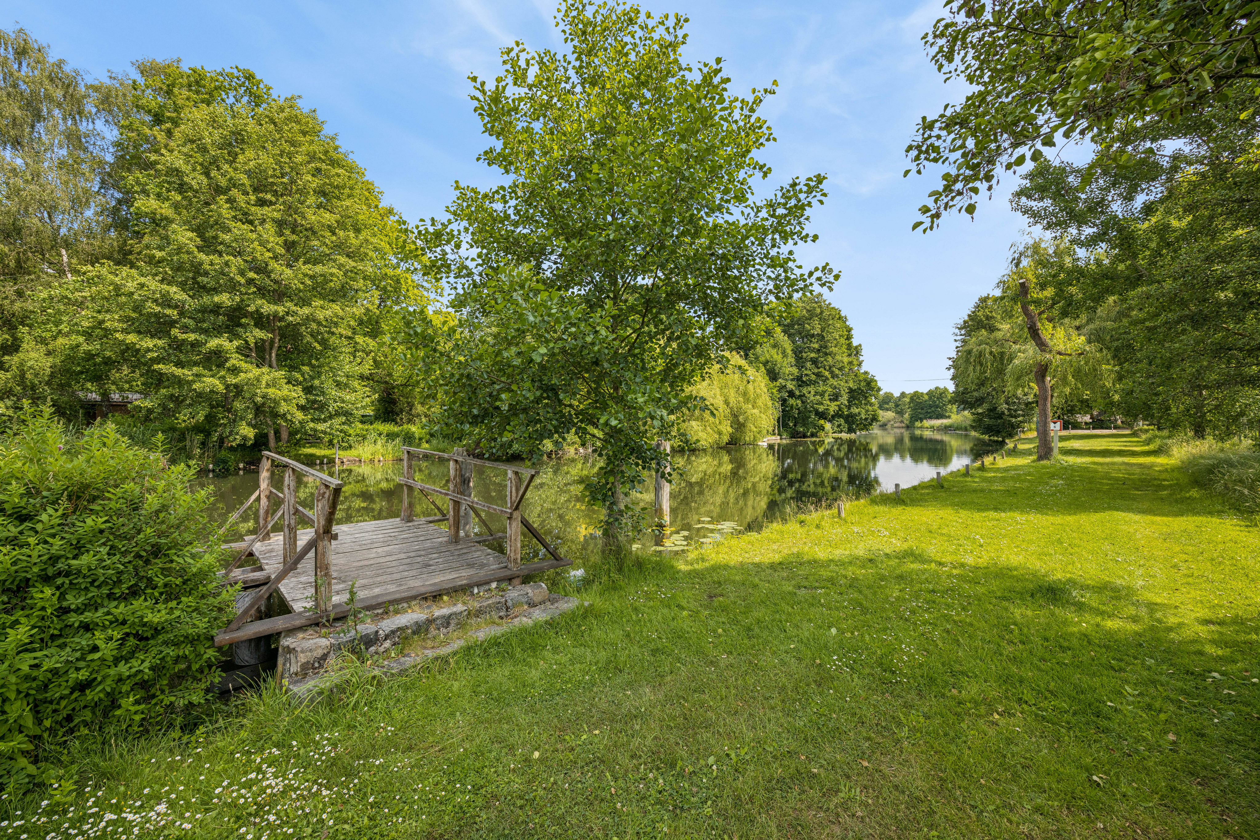 A wooden dock extends into a calm river surrounded by lush green trees and a grassy bank under a clear blue sky.