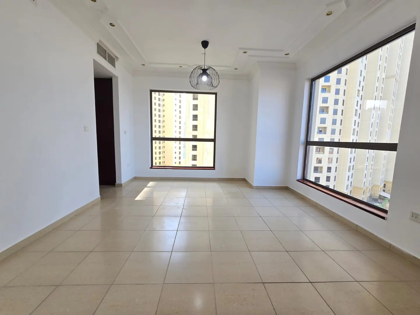 Bright, empty room with beige tile floor, white walls, and two large windows. A modern black light fixture hangs from the ceiling. High-rise buildings are visible outside.