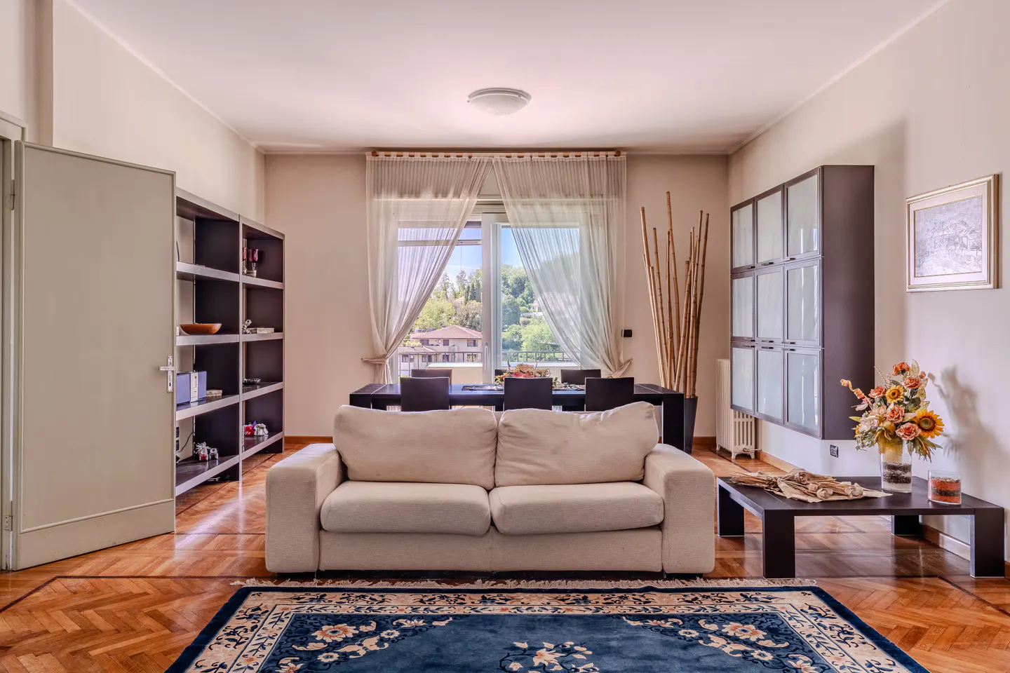 Living room with a white sofa, dark wood dining table and shelves, and a blue patterned rug on a herringbone wood floor.