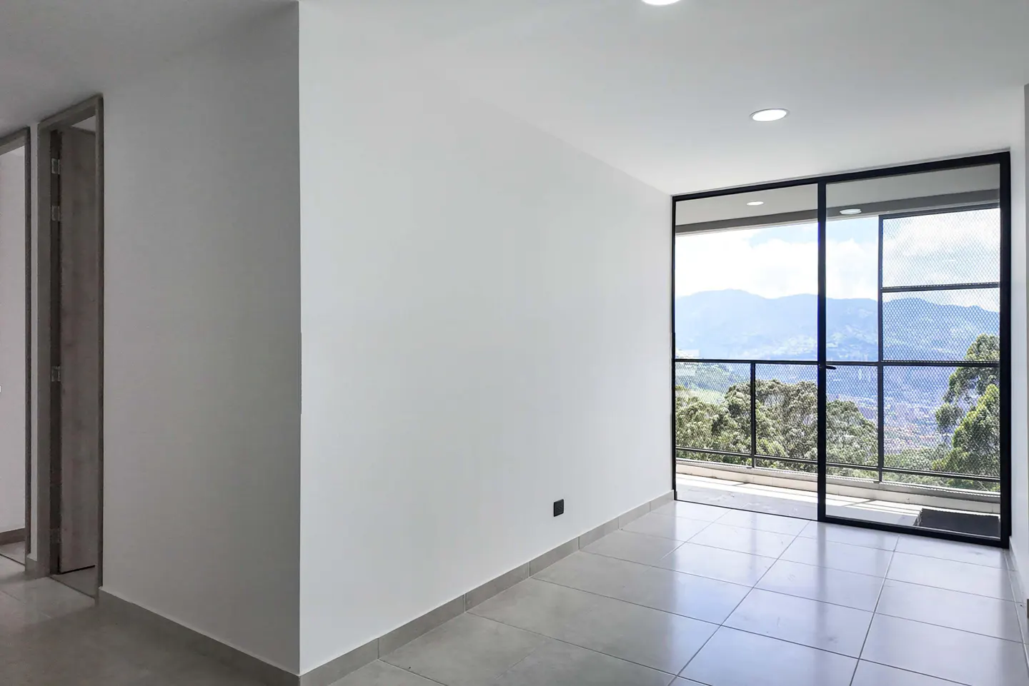 Bright, empty room with white walls and gray tile floor. A black-framed sliding glass door leads to a balcony with mountain views. A wood-look door is visible on the left.
