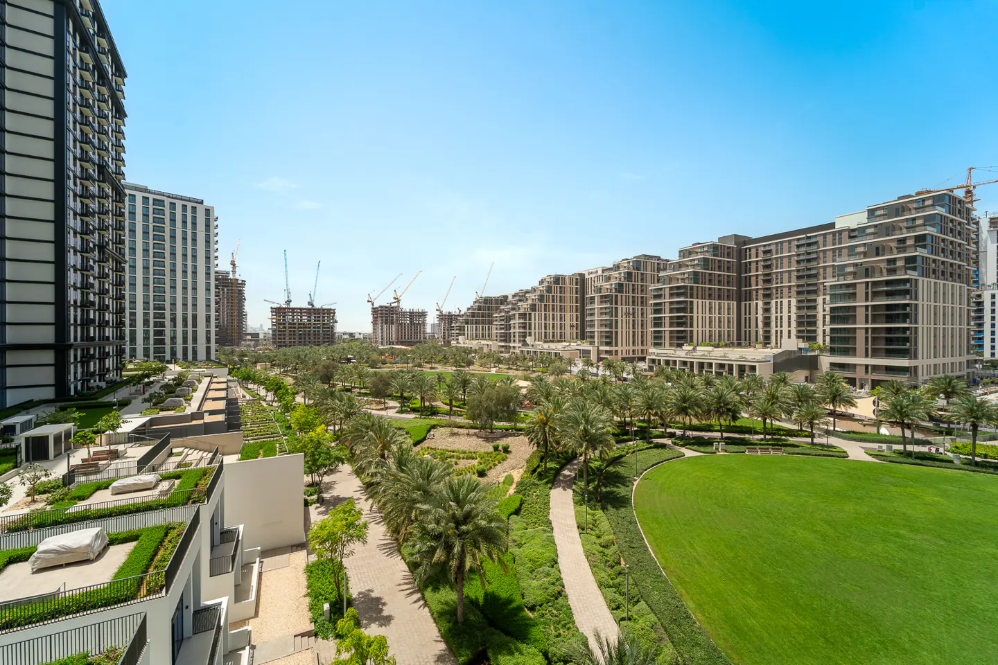 View of modern apartment buildings surrounding a green park with palm trees under a blue sky. Cranes are visible in the background.