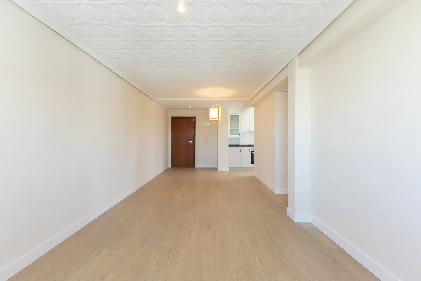 Empty apartment with light wood floors, white walls, and a textured white ceiling. A brown door and a glimpse of a white kitchen are visible.