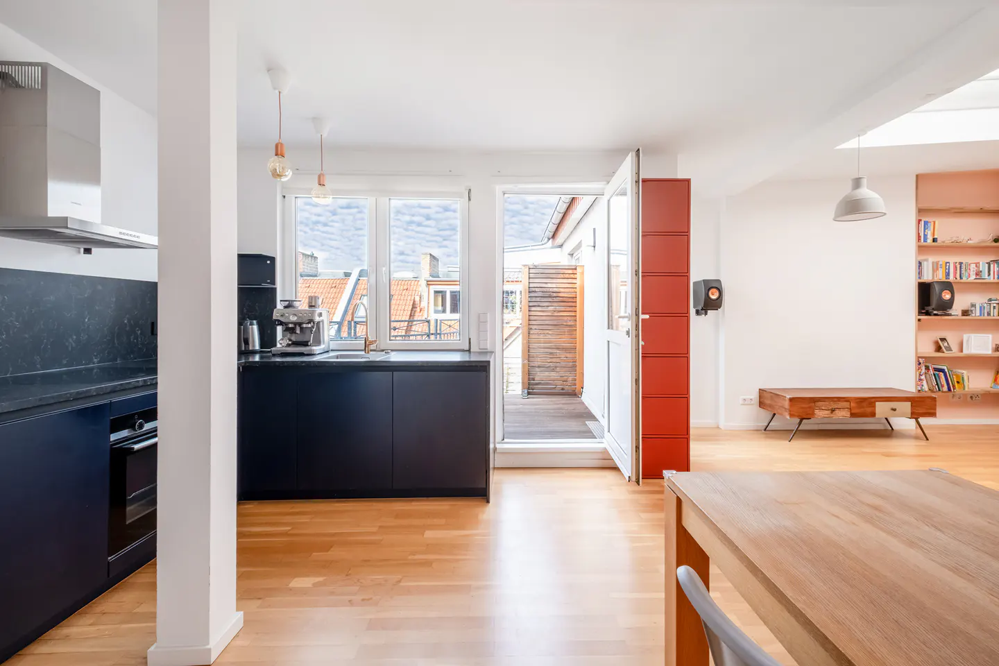 Bright, modern apartment interior with wood floors, black cabinets, and a red door leading to a balcony. A bookshelf and wooden table are visible.