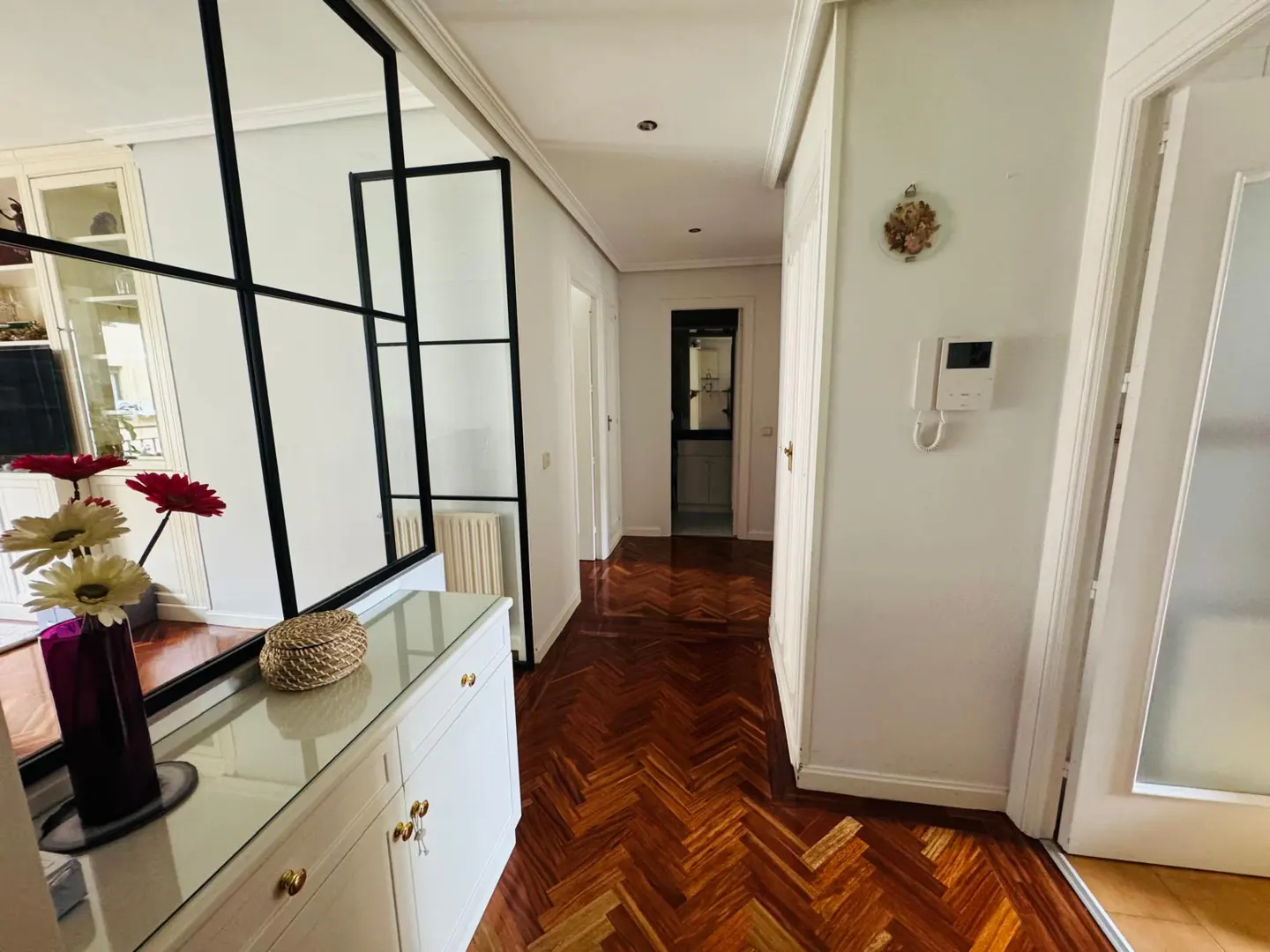 Hallway with herringbone wood floors, white walls, and black-framed glass partition. A white cabinet with flowers sits near the partition.
