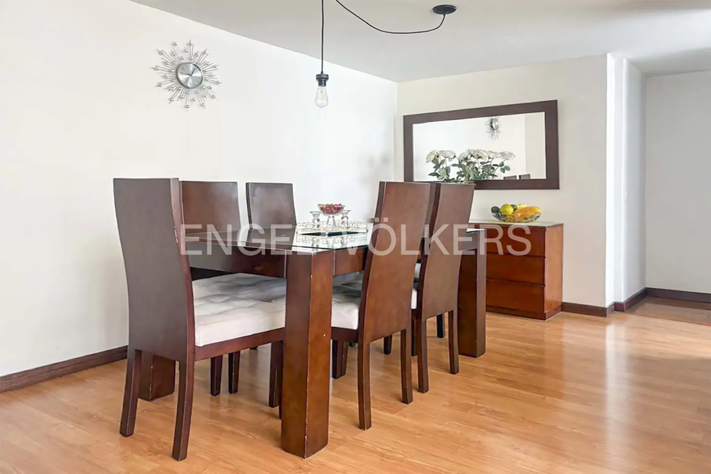 Dining room with a glass-topped table, six brown chairs, and a wooden floor. A mirror and a wooden dresser are in the background.