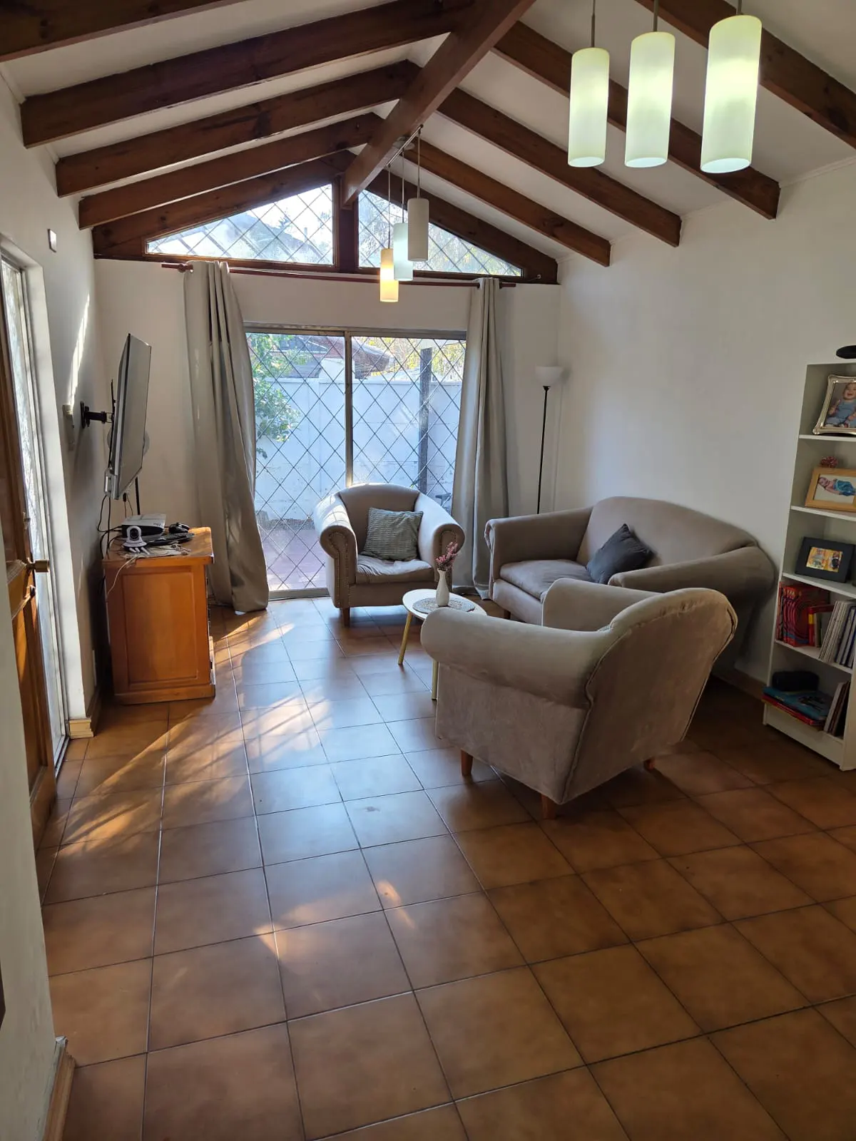 Living room with vaulted wood-beam ceiling, tile floors, beige sofas, and large windows with lattice security bars.