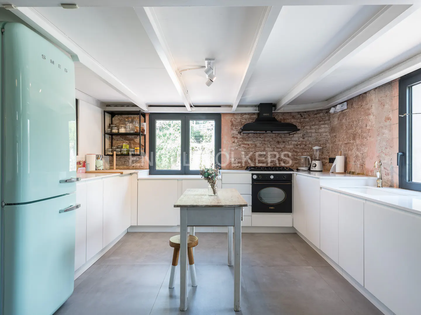 Bright kitchen with white cabinets, a light blue SMEG refrigerator, and a black oven. A small table and stool sit in the center.