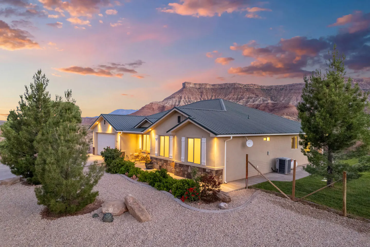 A tan house with a green roof sits in front of a red rock mountain at sunset.