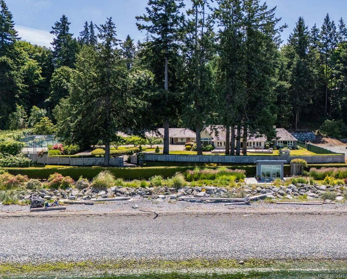 A waterfront property with a large house, green trees, and a rocky beach. The house is partially hidden by the trees.