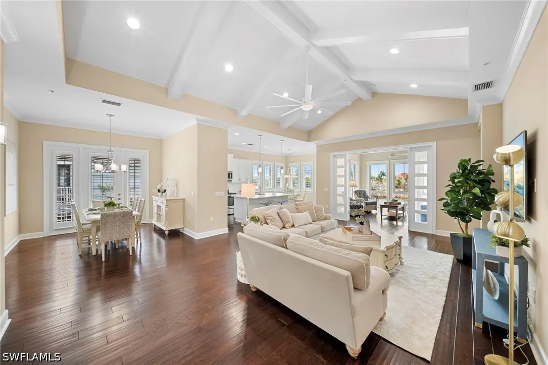 Bright, open-concept living space with dark wood floors, cream sofas, and a dining area. Vaulted ceilings with white beams and a ceiling fan.