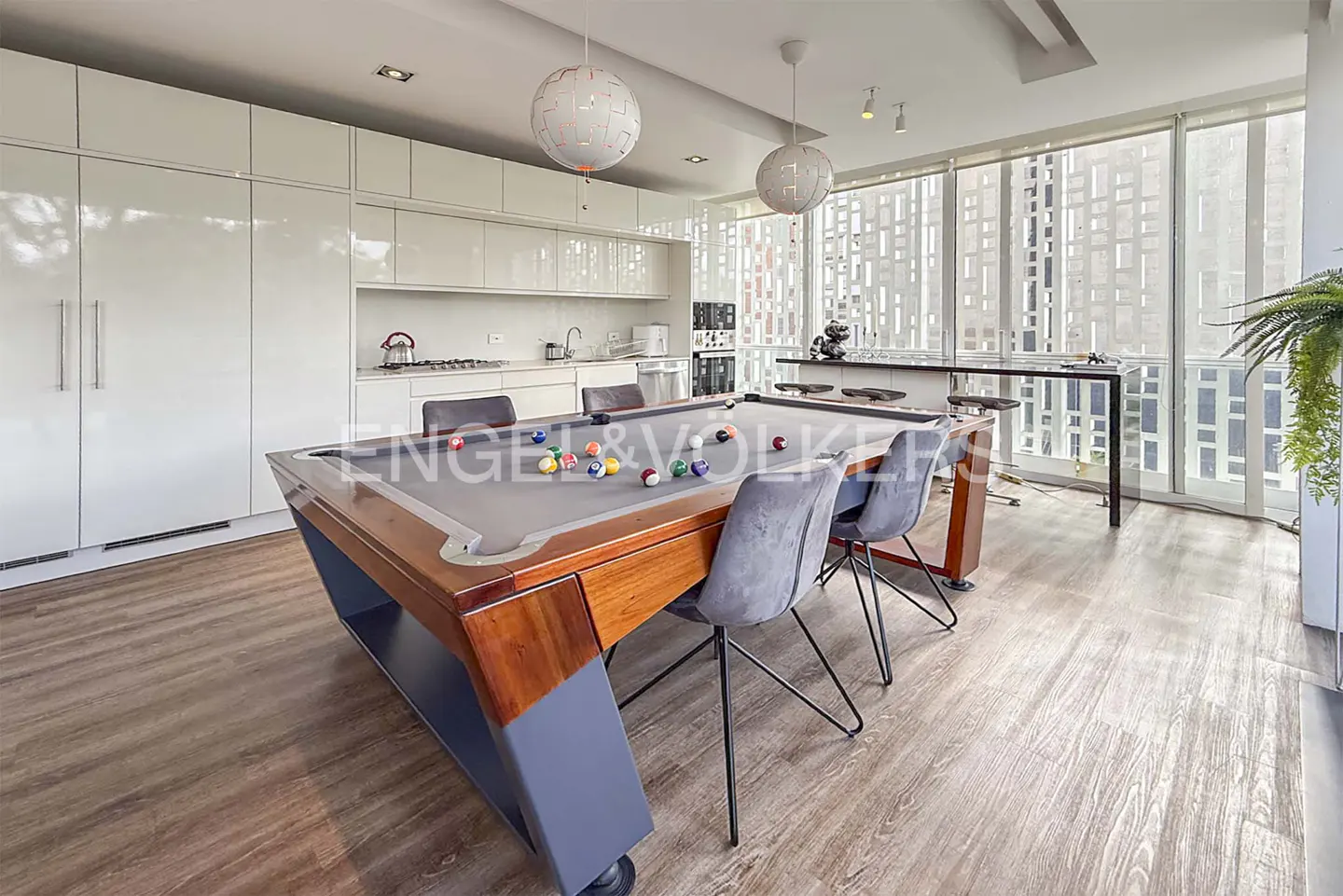 A modern, open-concept room with a pool table, kitchen, and bar area. The pool table is wood with gray felt, and the kitchen has white cabinets.