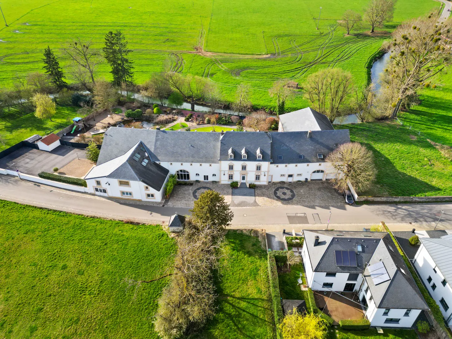 Aerial view of a long, white, multi-sectioned house with a gray roof, surrounded by green fields and trees.