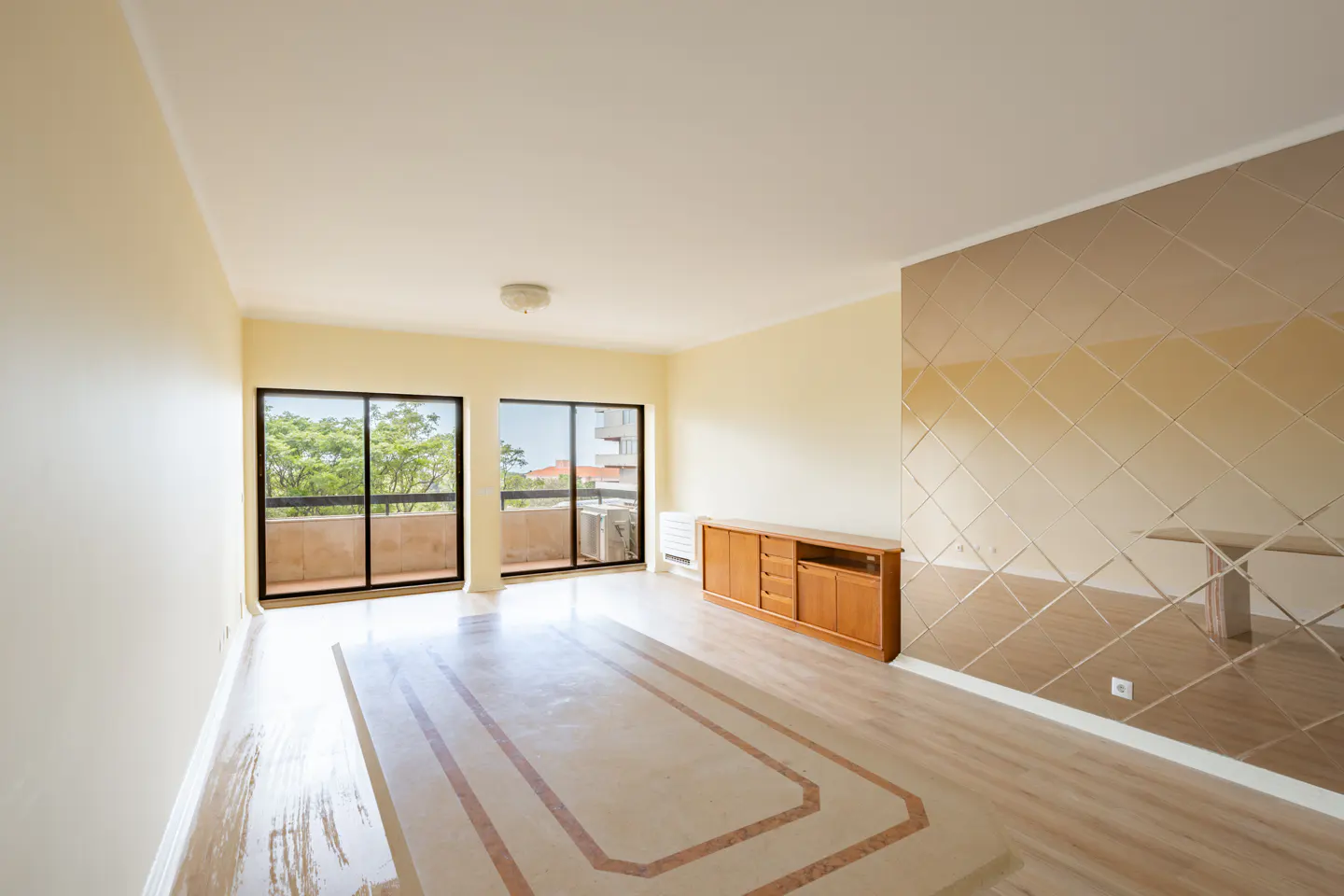 Bright, empty living room with beige walls, light wood floors, and a mirrored accent wall. Two black-framed sliding doors lead to a balcony. A wooden cabinet sits against the wall.