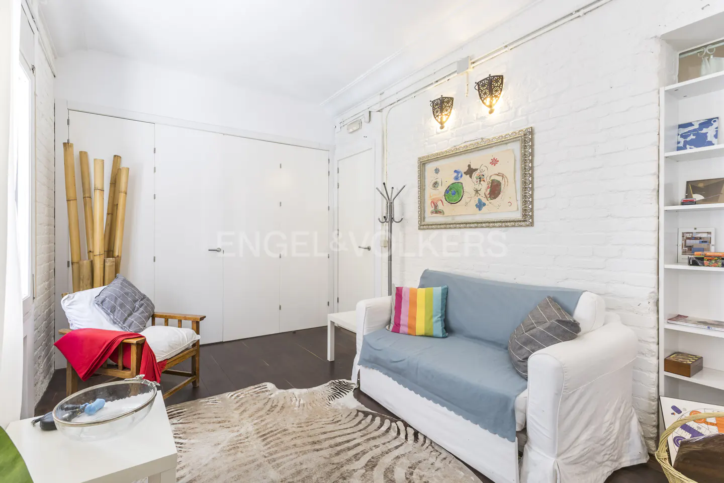 Bright living room with white brick walls, a blue sofa with a rainbow pillow, and a zebra-print rug. A wooden chair sits near bamboo stalks.