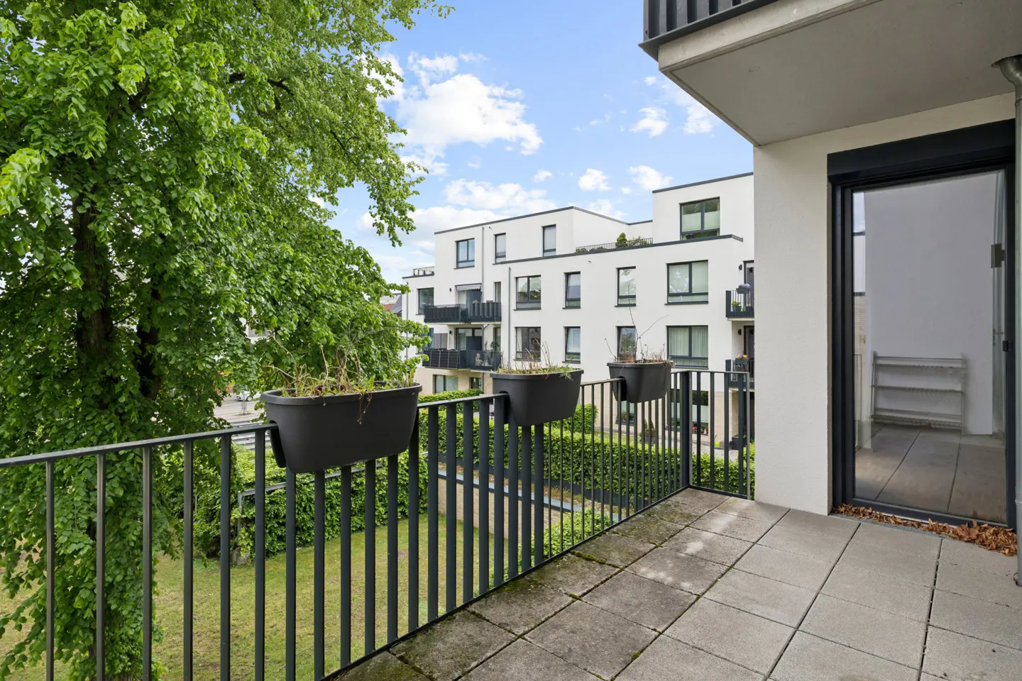 Balcony view with black railing, planters, and gray tiled floor. White apartment buildings and green trees in the background.