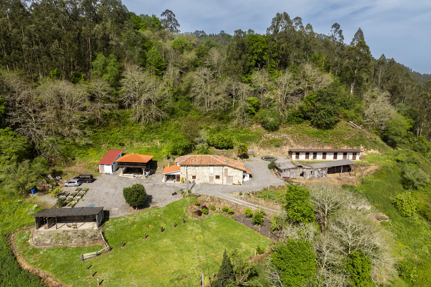 Aerial view of a stone house with a red tile roof, surrounded by green grass and trees on a hillside.