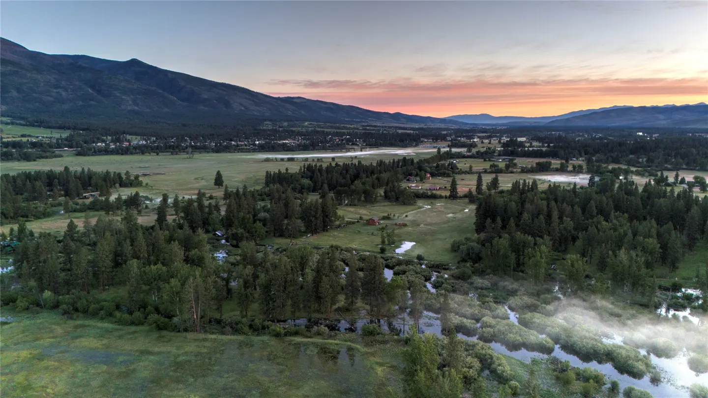 Aerial view of a valley with trees, a river with fog, and mountains in the background at sunset.
