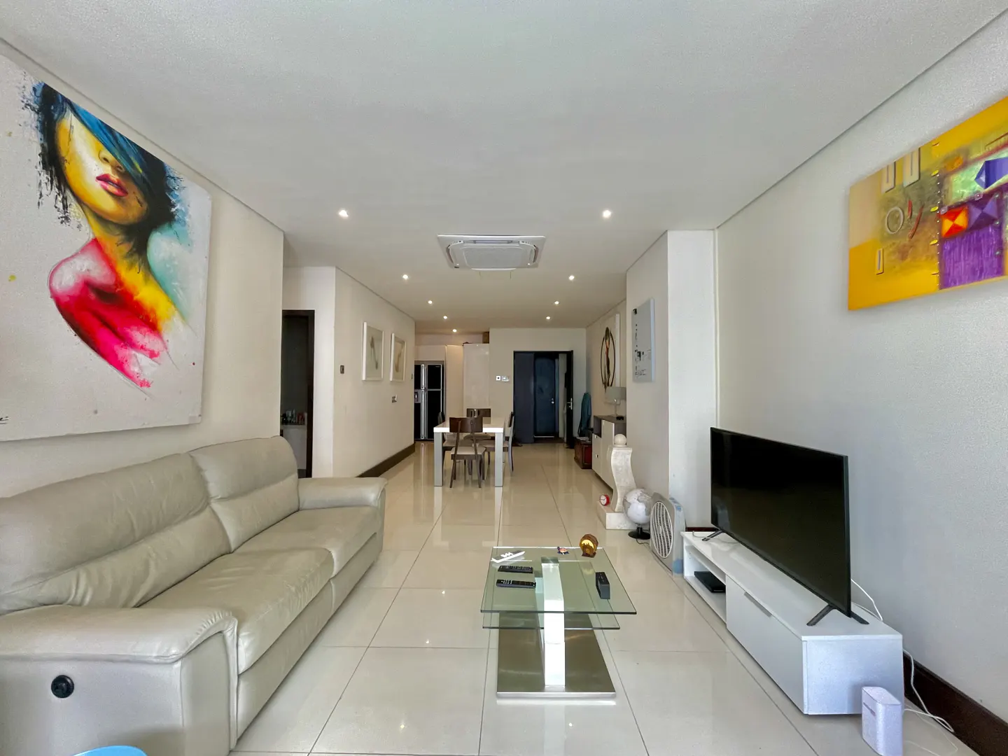 Bright, modern living room with white tile floors, a cream sofa, and a glass coffee table. Colorful art adorns the walls.
