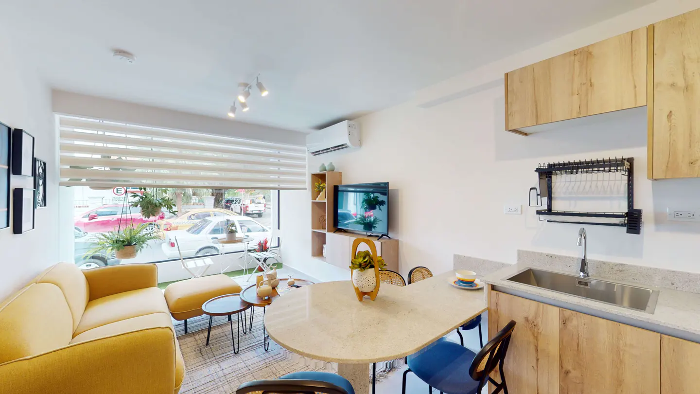 Bright, modern apartment interior with a yellow sofa, round table with blue chairs, and wood cabinets. Large window with blinds lets in natural light.
