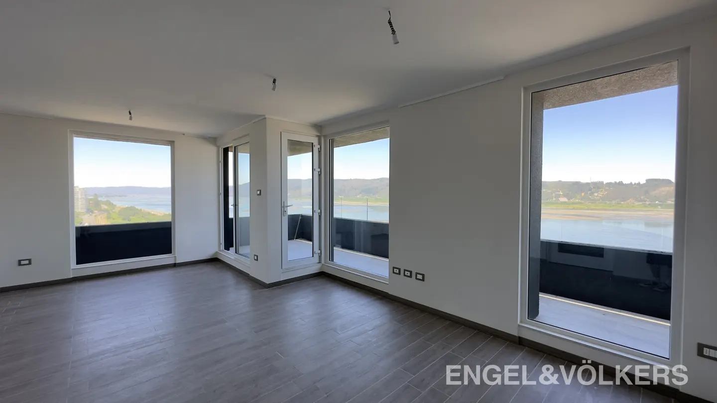 A bright, empty room with gray floors and white walls. Large windows offer a view of a body of water and distant land. Engel & Völkers logo in the corner.