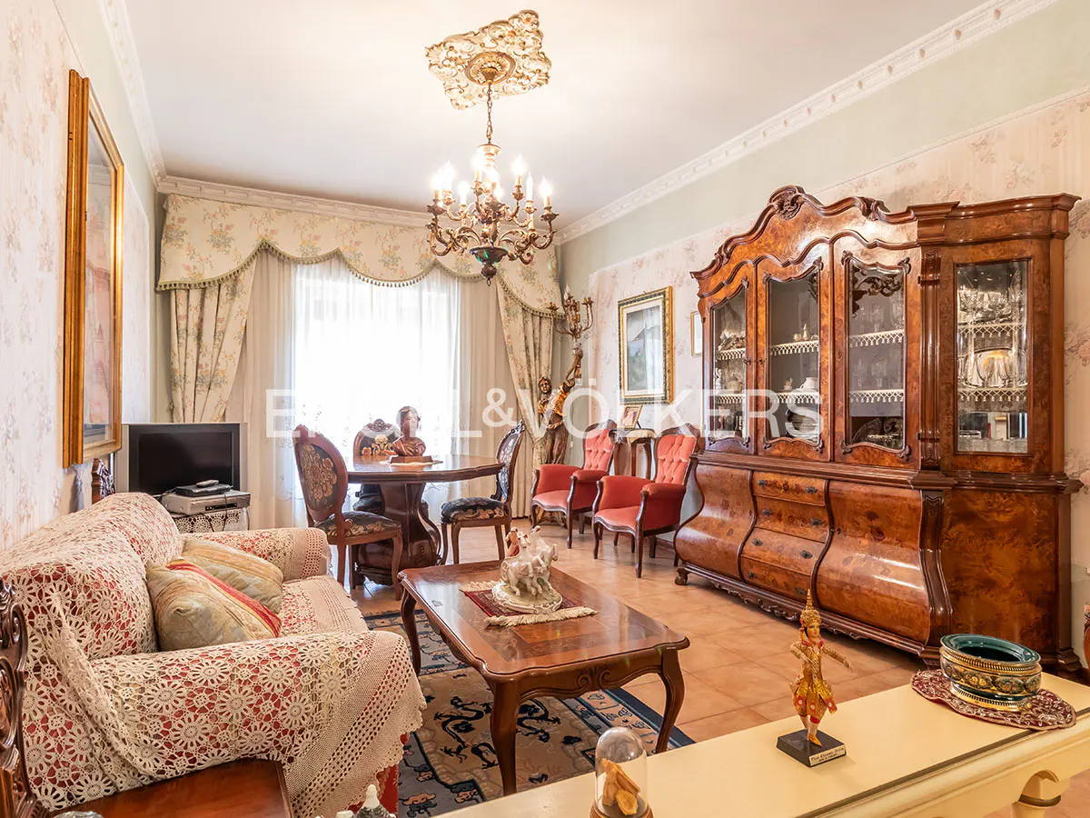 Ornate living room with floral wallpaper, a chandelier, and antique furniture including a large wooden cabinet and a sofa with a lace cover.