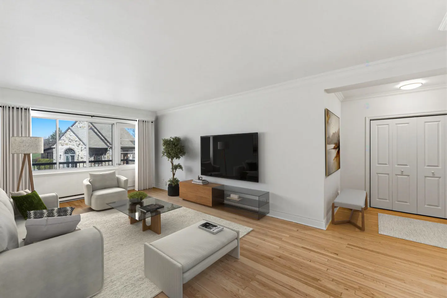 Bright living room with hardwood floors, white walls, and large windows. Furniture includes a sofa, chairs, glass coffee table, and TV.
