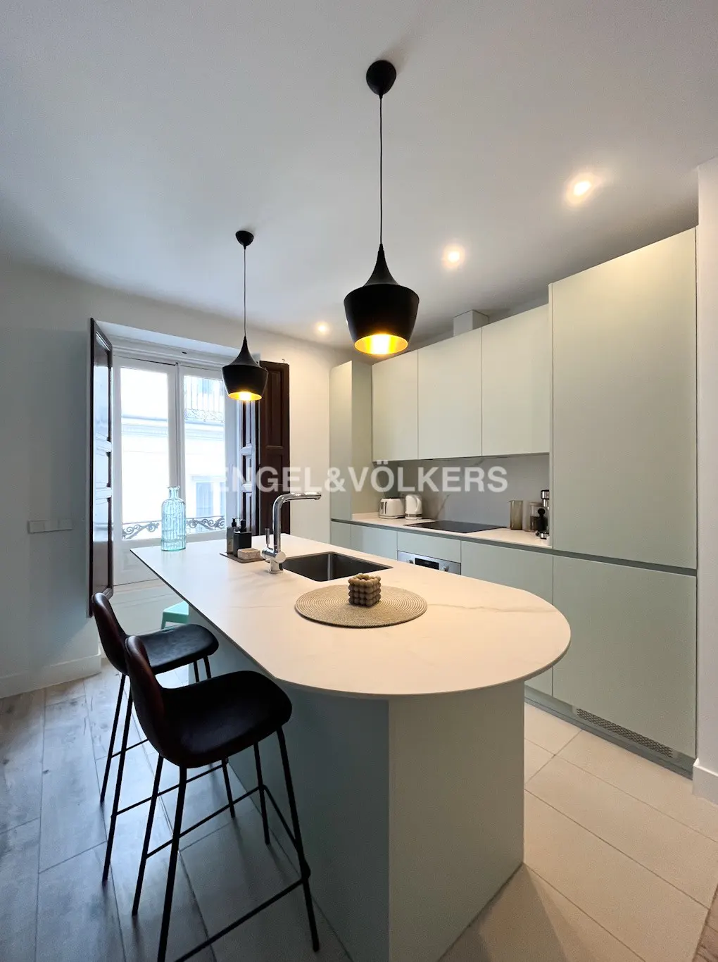 Modern kitchen with a large island, black bar stools, and pendant lights. Light green cabinets and white countertops create a clean, airy feel.