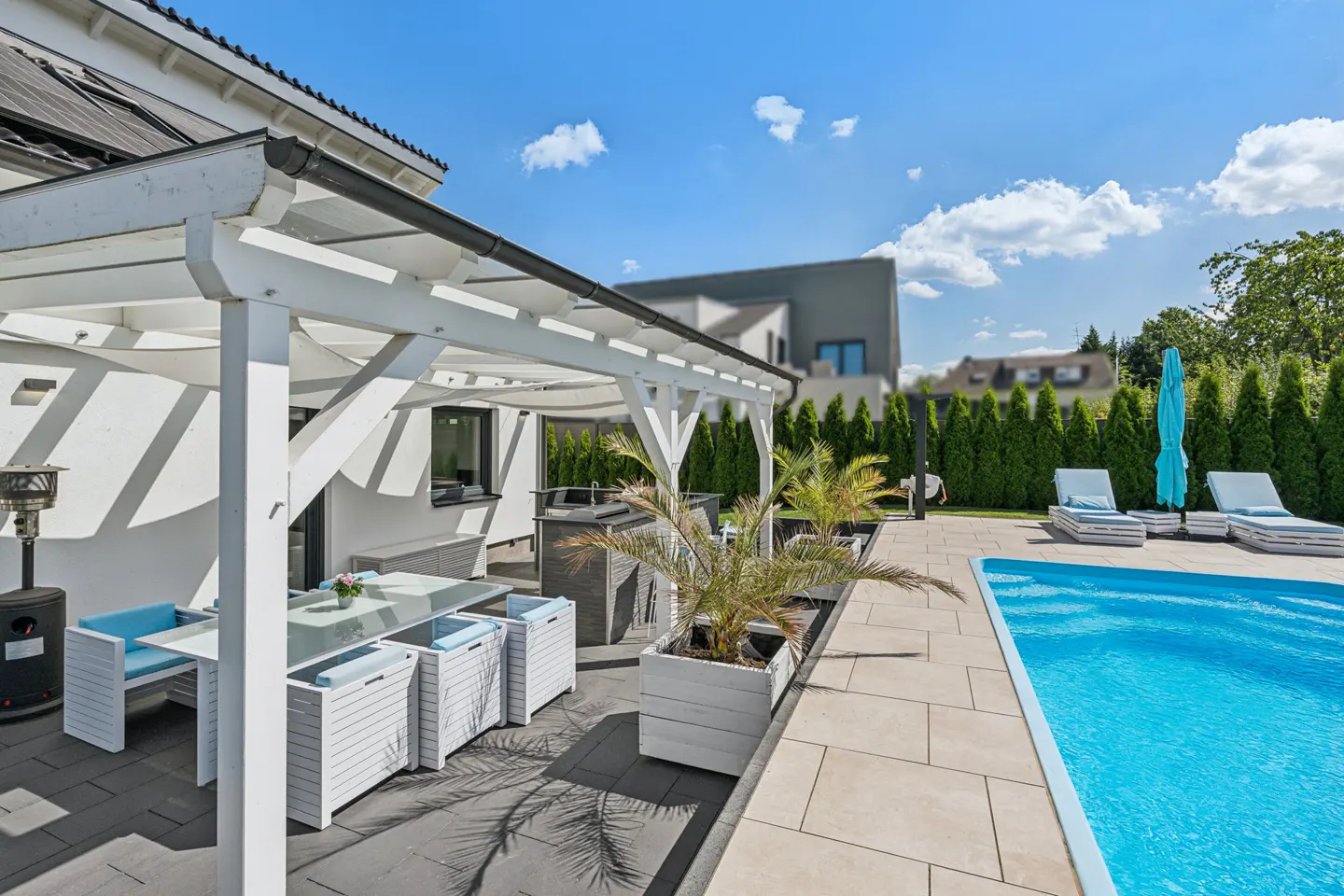 Outdoor patio with a white pergola, table, chairs, and a pool with lounge chairs on a sunny day.