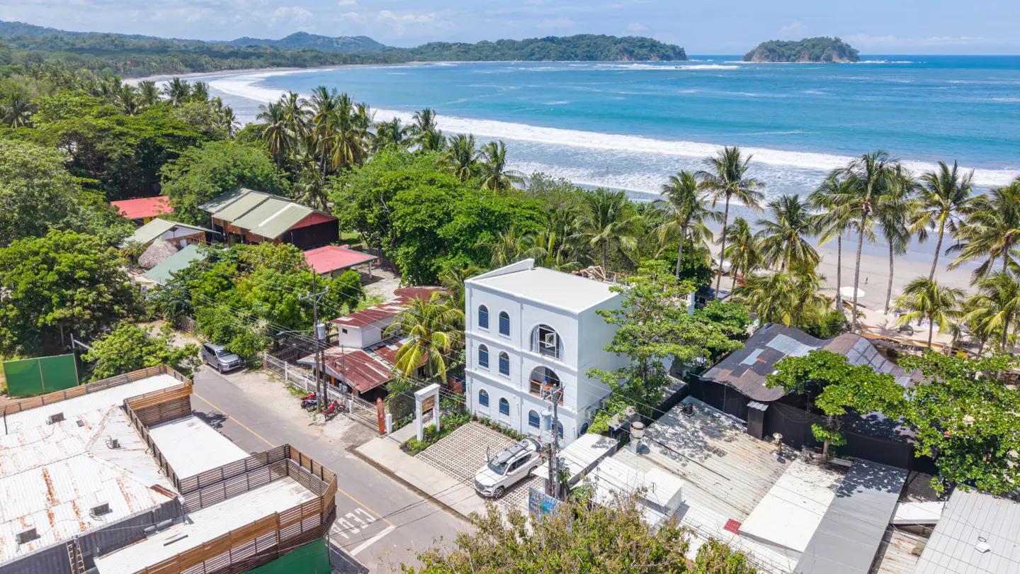 Aerial view of a white three-story building near a tropical beach with palm trees, blue ocean, and surrounding houses.