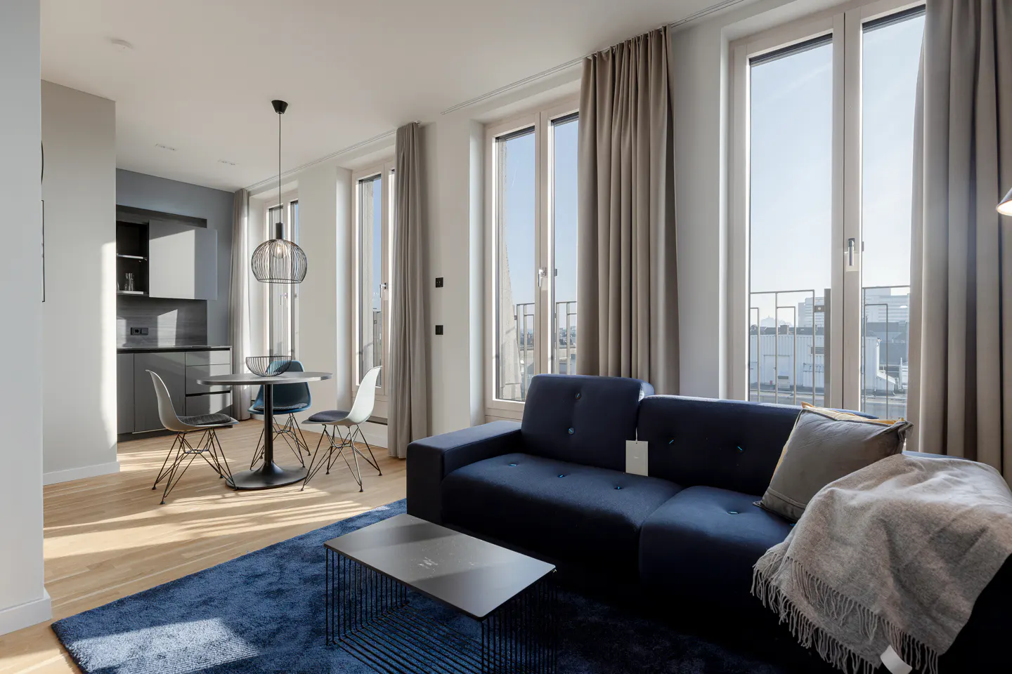 Bright apartment interior with a blue sofa, rug, and a table with chairs near a gray kitchen. Tall windows with beige curtains.