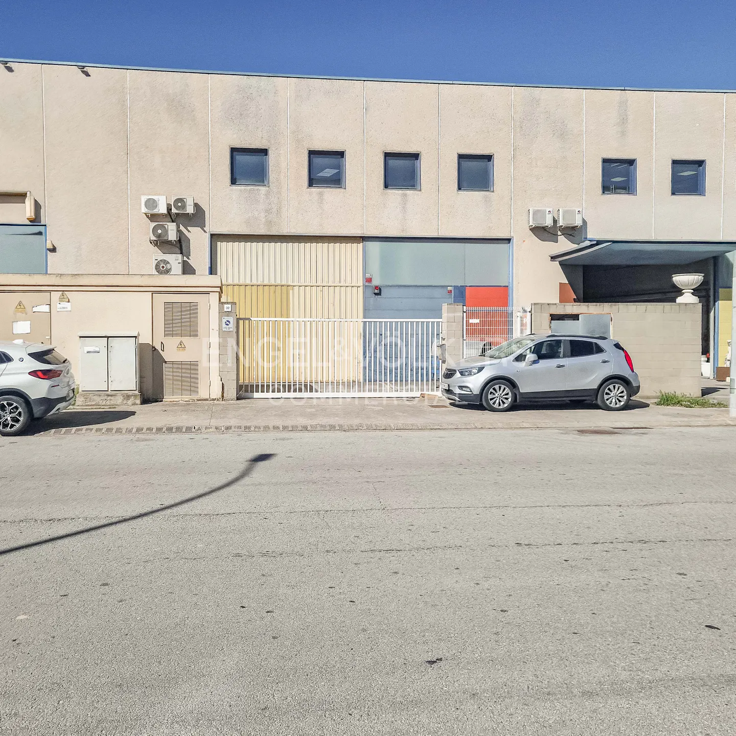 A beige industrial building with a white gate and a silver car parked in front.