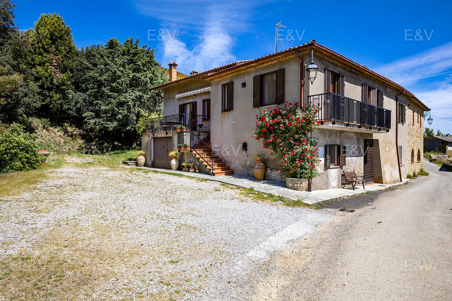 Exterior of a two-story house with a gravel driveway and a red rose bush. The house has a balcony and a stone facade.