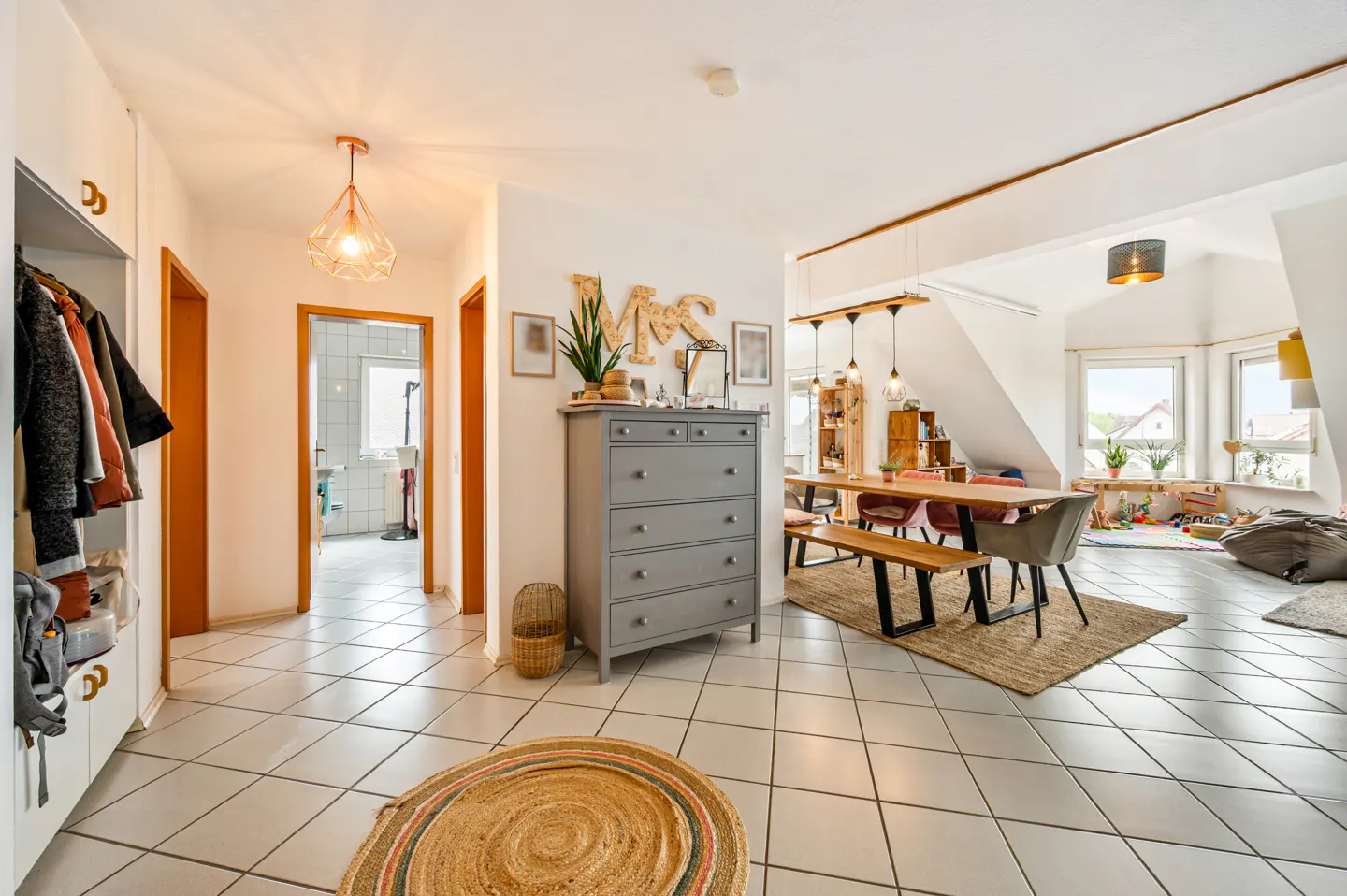 Bright open-plan home with tiled floors, a grey dresser, a wooden dining table, and a view into a sunlit living area.