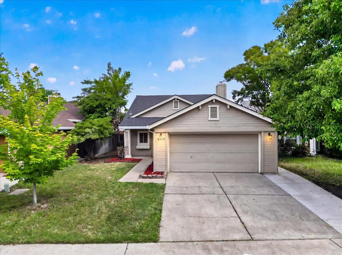 Beige single-story house with a gray roof, two-car garage, concrete driveway, and green lawn under a blue sky.