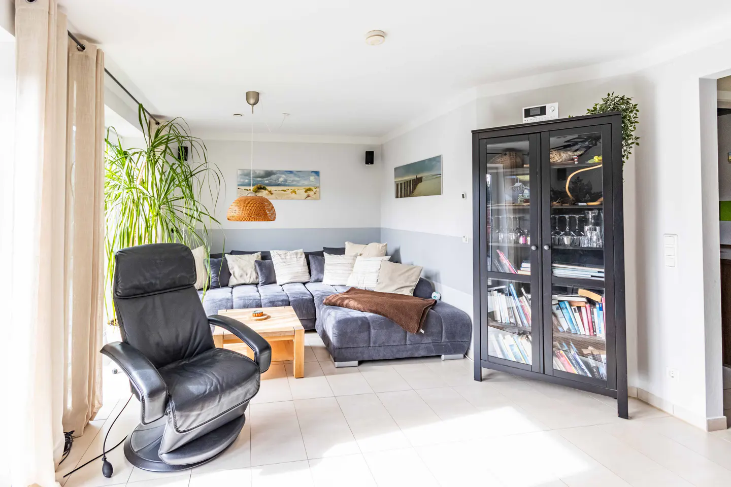 Bright living room with a gray sectional sofa, black leather recliner, and a black glass-door cabinet filled with books and glassware.
