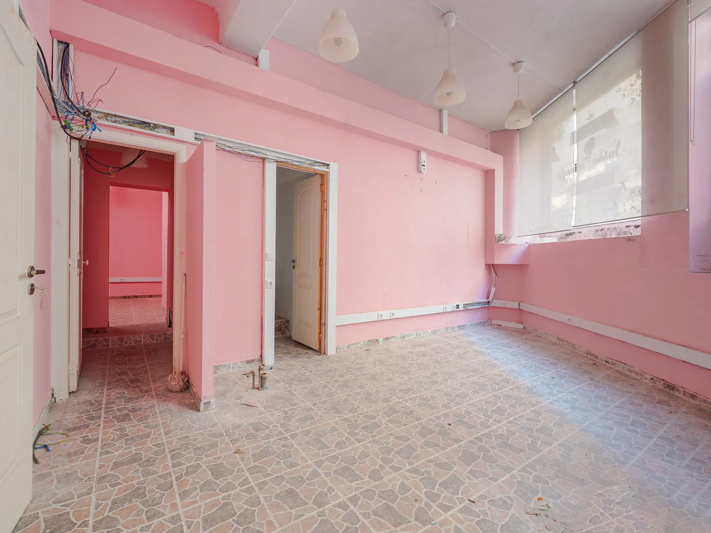 An empty pink room with tile floors, white trim, and three pendant lights. There are three open doorways and a window with a shade.