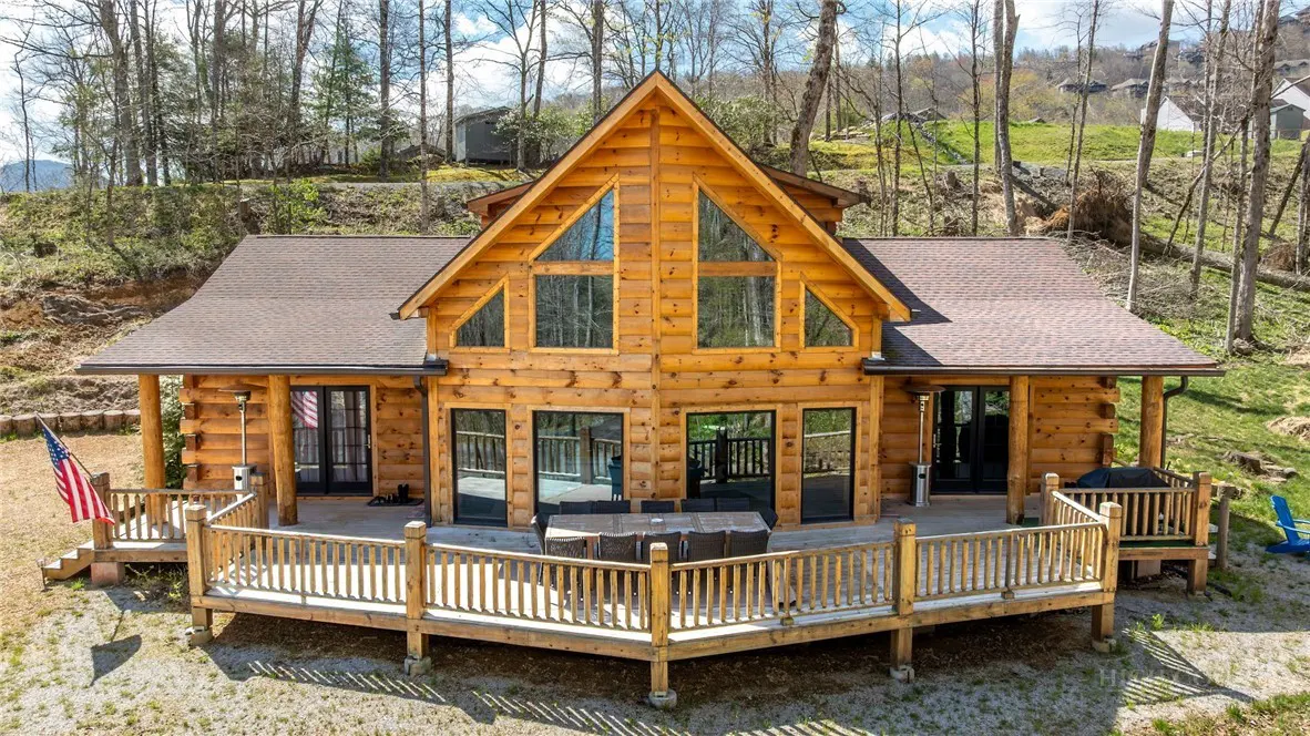 A rustic log cabin with a large wooden deck, outdoor dining set, and American flag, set against a wooded mountain backdrop.